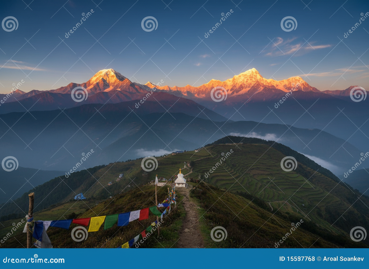 Snow-capped Himalayan peaks at golden hour above green terraced hills, with colourful prayer flags in the foreground