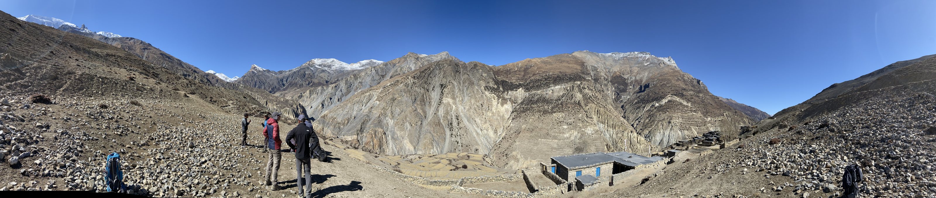 Panorama of a Himalayan valley in Nepal: snow-capped peaks, steep mountainsides, trekkers on a rocky trail and a stone house with blue windows
